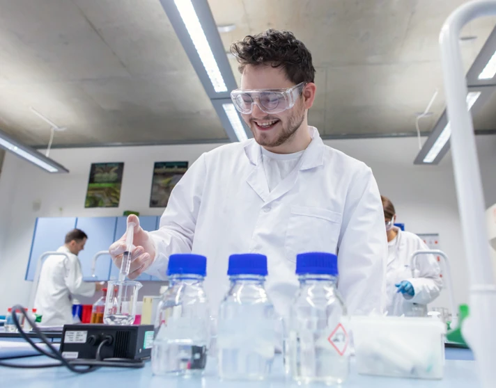 Male student wearing lab coat and goggles pipetting liquid during science experiment Male student wearing lab coat and goggles pipetting liquid during science experiment